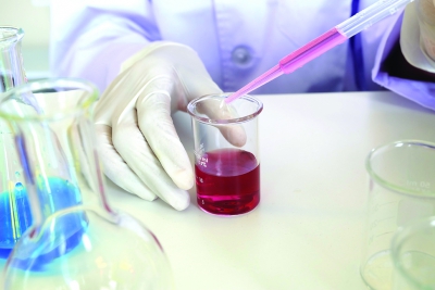 A scientist holding a pipette and a beaker containing a pink liquid on a white table