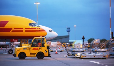 East Midlands Airport, a yellow DHL branded cart pulls along plane equipment, behind it a yellow plane and a white plane are pictured