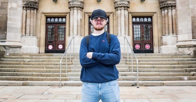 student wearing black cap stands with arms crossed outside a pillar fronted hall with set of stairs leading up