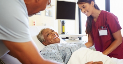 two smiling medical professionals stand over the bed of a lady who is smiling up at them