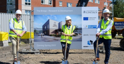 three people with hardhats rest their foot on a spade and smile at the bridge research and development