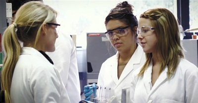 3 female scientists in googles and white lab coats talking around some test tubes
